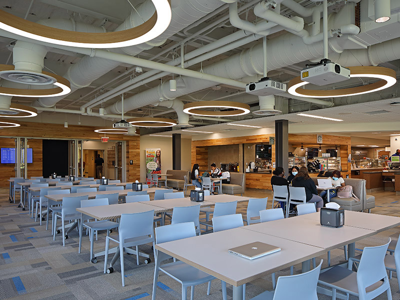 Modern dining and student commons area at Penn State Lehigh Valley featuring open seating, circular LED light fixtures, exposed ductwork, and wood-accent finishes
