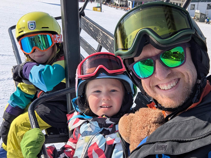 A photo of David Rosenberger and two children wearing helmets and ski goggles ride a ski lift, dressed in colorful winter apparel with a snow-covered mountain landscape in the background.