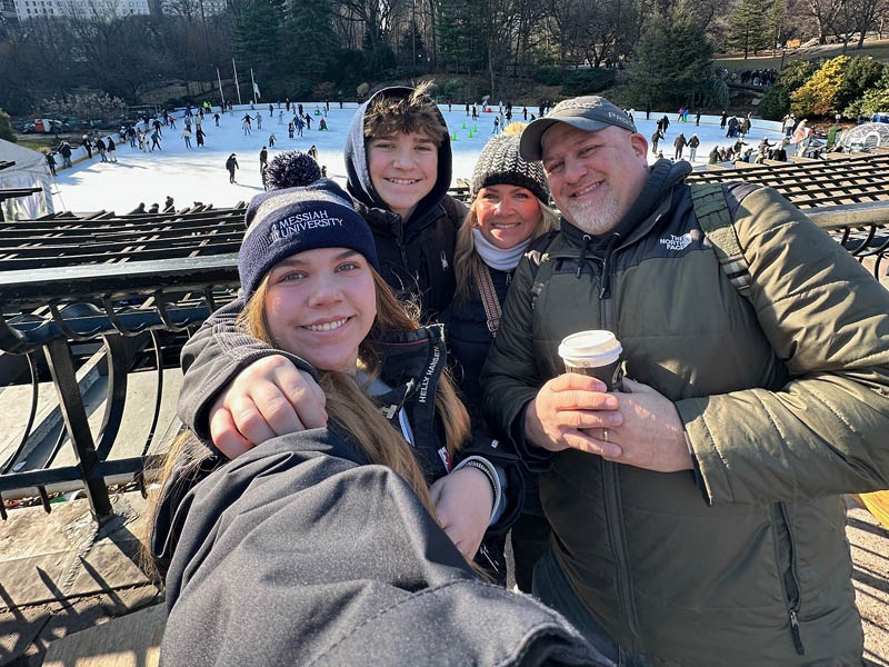 Jason and his family wearing winter coats and hats smile for a selfie in front of a large outdoor ice-skating rink, with numerous skaters and trees visible in the background.