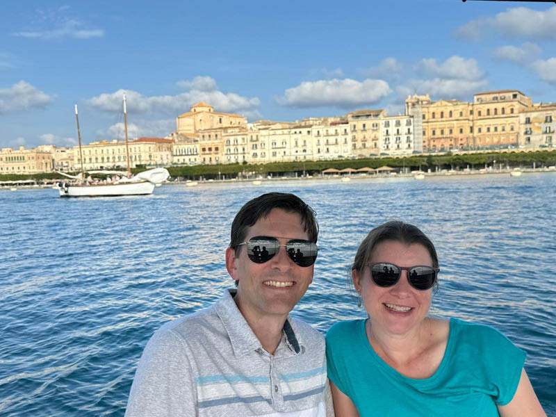 Benjamin D. Basom and his wife smiling in sunglasses while taking a waterfront selfie, with a scenic coastal city, historic buildings, and boats in the background under a bright blue sky.