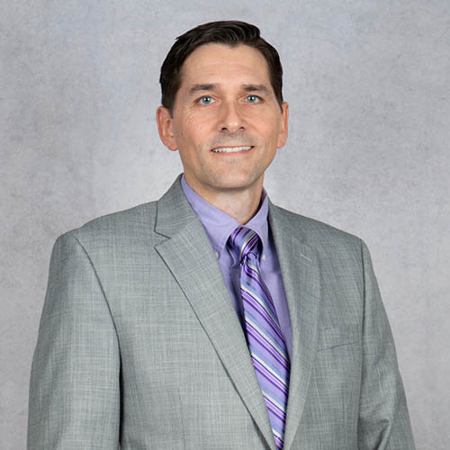 Benjamin D. Basom professional headshot in a gray suit and purple shirt with a striped tie, smiling against a neutral studio background.