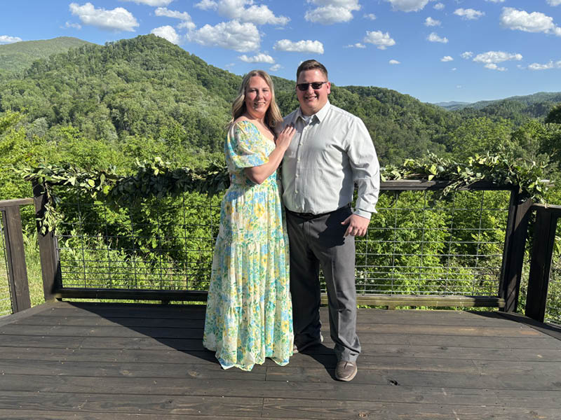 Connor D. Smith and his fiancée smiling together on a wooden deck overlooking lush green mountains, with a scenic landscape and blue sky in the background.