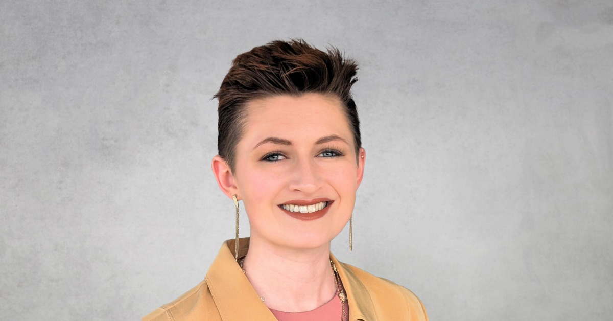 Professional headshot of Sarah Halstead with short styled hair, smiling, wearing a tan jacket and earrings against a neutral gray background