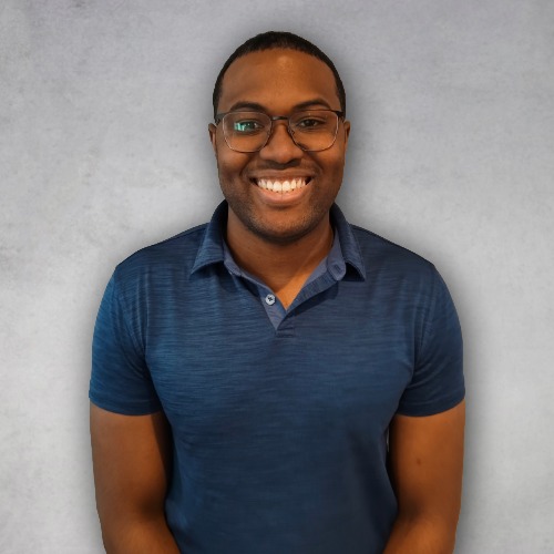 Professional headshot of Ethan Brown wearing glasses and a navy blue polo shirt, smiling against a gray studio background.
