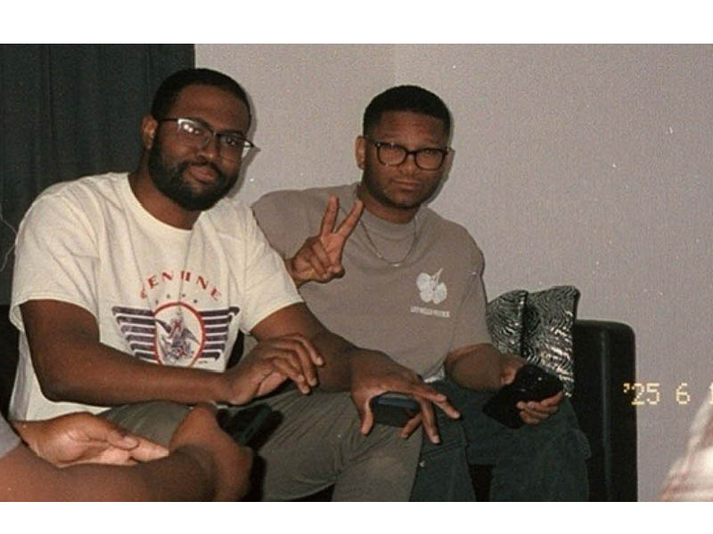 Ethan Brown wearing a white graphic T-shirt and glasses, sitting indoors with friends in a casual social setting.