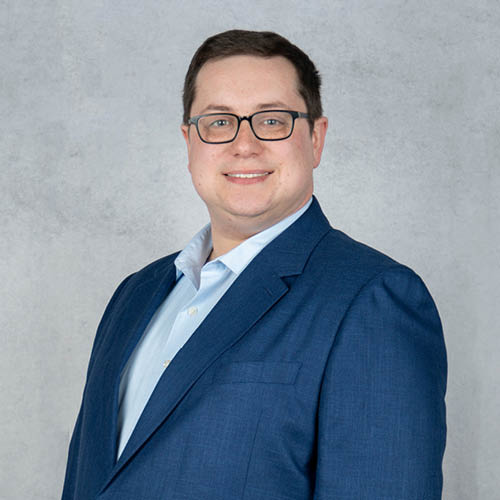 Professional headshot of Michael Duffy wearing a blue suit jacket and light blue dress shirt, smiling against a gray studio background.