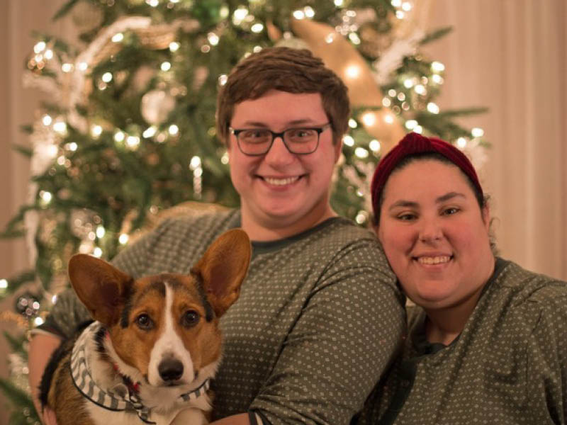 Michael Duffy smiling with his partner and their corgi in matching pajamas in front of a decorated Christmas tree.