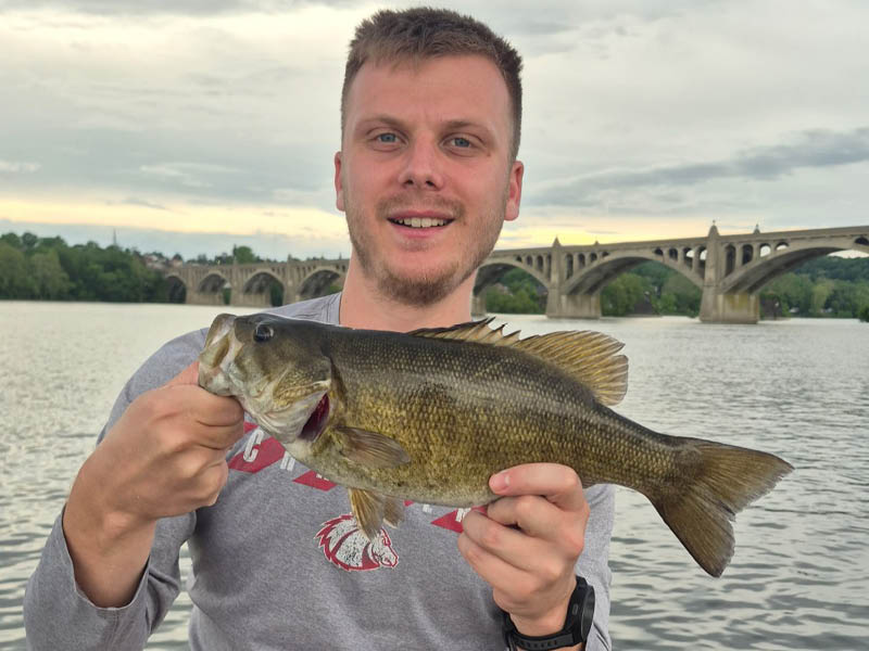 Caleb holding a largemouth bass while standing by a river with a bridge in the background.