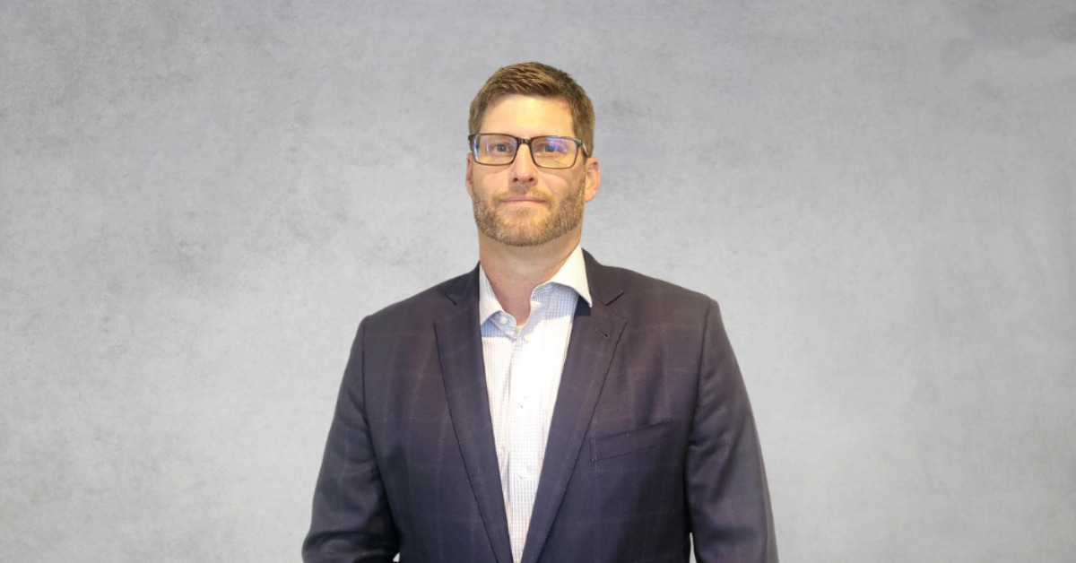 Isaac K. Sluga smiles in a professional headshot while wearing a navy suit jacket, light blue dress shirt, and glasses against a neutral gray background.