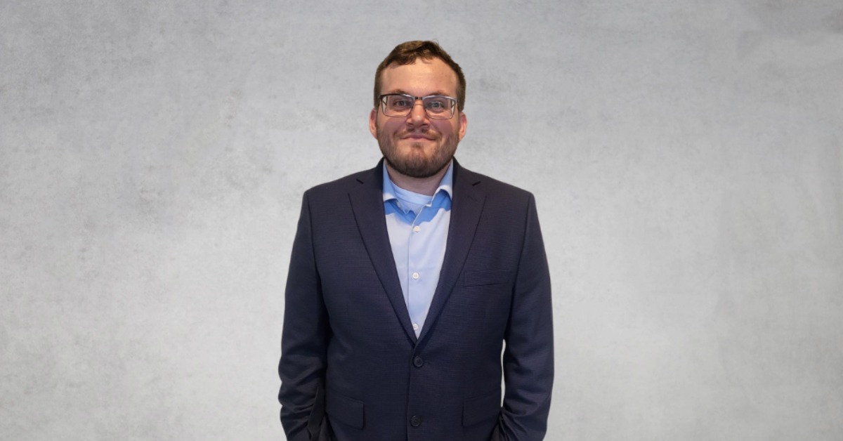 Matthew F. Pizano smiles in a professional headshot while wearing a navy blazer, light blue dress shirt, and glasses against a neutral gray background.