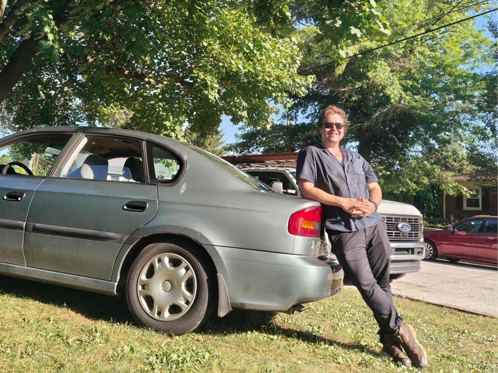 Samuel wearing sunglasses leaning against a parked car outdoors on a sunny, tree-lined residential street.